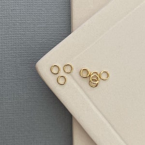 May include: A close-up of a group of small, gold jump rings on a white surface. The jump rings are arranged in a loose cluster, with some overlapping.