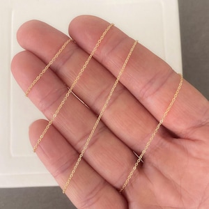May include: A delicate gold-coloured chain necklace, displayed on a hand. The chain is thin and appears to be made of small, connected links. The background is a neutral grey and white.