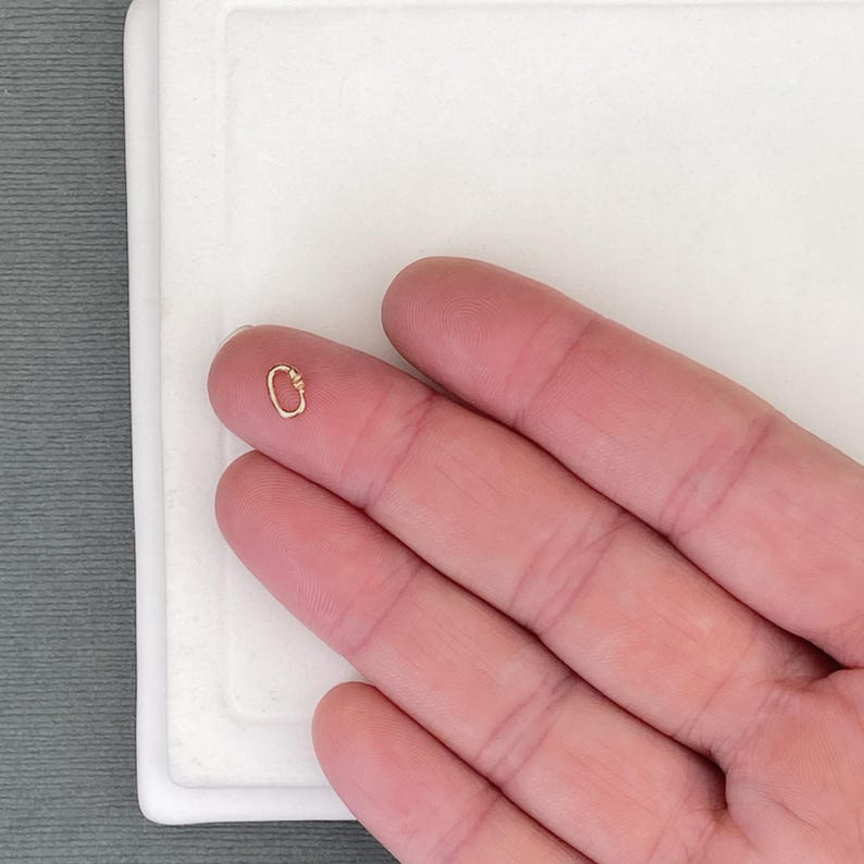 May include: A small, oval-shaped gold-toned metal spring ring clasp featuring a hinged mechanism. The jewellery component is metallic with a polished finish and is shown resting on a fingertip to demonstrate its diminutive scale and size.