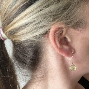 May include: A pair of gold dangle earrings with a small, round, textured charm. The charm has a design that resembles a stylized flower or star.