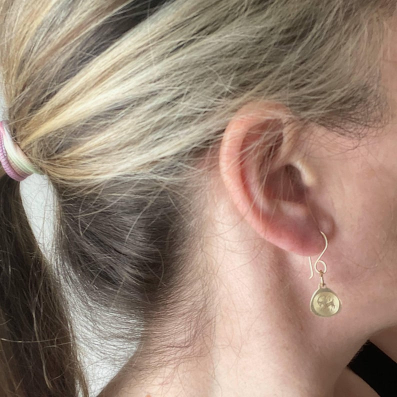 May include: A pair of gold dangle earrings with a small, round, textured charm. The charm has a design that resembles a stylized flower or star.