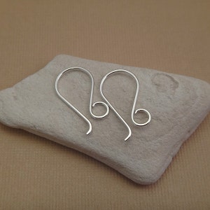 May include: Two silver earring hooks with a small loop at the top. The hooks are on a light brown surface.