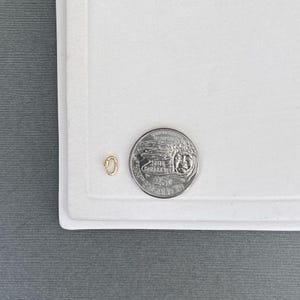 May include: A small gold-toned oval jump ring or connector link positioned next to a United States quarter coin for scale. The metal link features a smooth, polished surface and a simple, continuous loop design, providing a clear size reference.