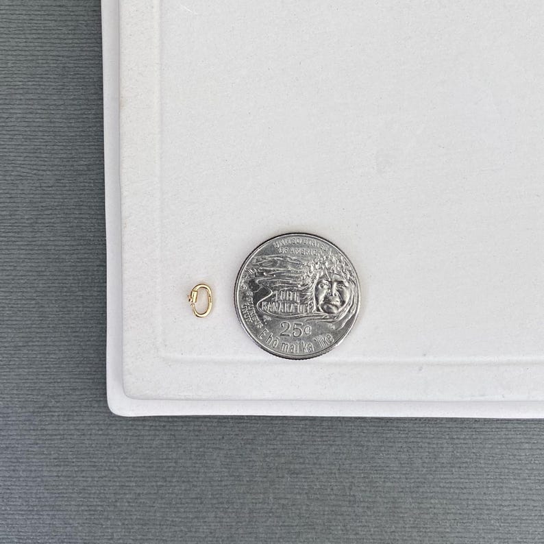 May include: A small gold-toned oval jump ring or connector link positioned next to a United States quarter coin for scale. The metal link features a smooth, polished surface and a simple, continuous loop design, providing a clear size reference.