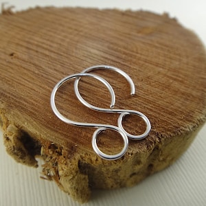 Puede incluir: Un pendiente plateado con un diseño abstracto único. El pendiente presenta una parte superior curvada y dos elementos circulares conectados, creando una forma distintiva. La joya se exhibe sobre una superficie de madera natural.