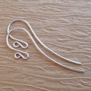 May include: Two silver earring hooks. Each hook features a looped design at the top and a long, slender, curved body. The earrings are set against a textured, light brown background.