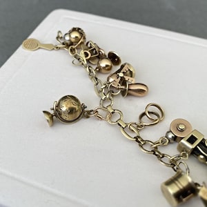 May include: A gold-tone charm bracelet featuring various dangling charms including a miniature globe, a tennis racket, a bell, and a small bucket. The bracelet is constructed from a series of interconnected oval links with a polished metallic finish.