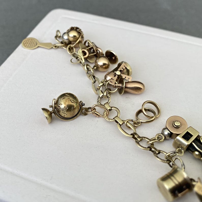 May include: A gold-tone charm bracelet featuring various dangling charms including a miniature globe, a tennis racket, a bell, and a small bucket. The bracelet is constructed from a series of interconnected oval links with a polished metallic finish.