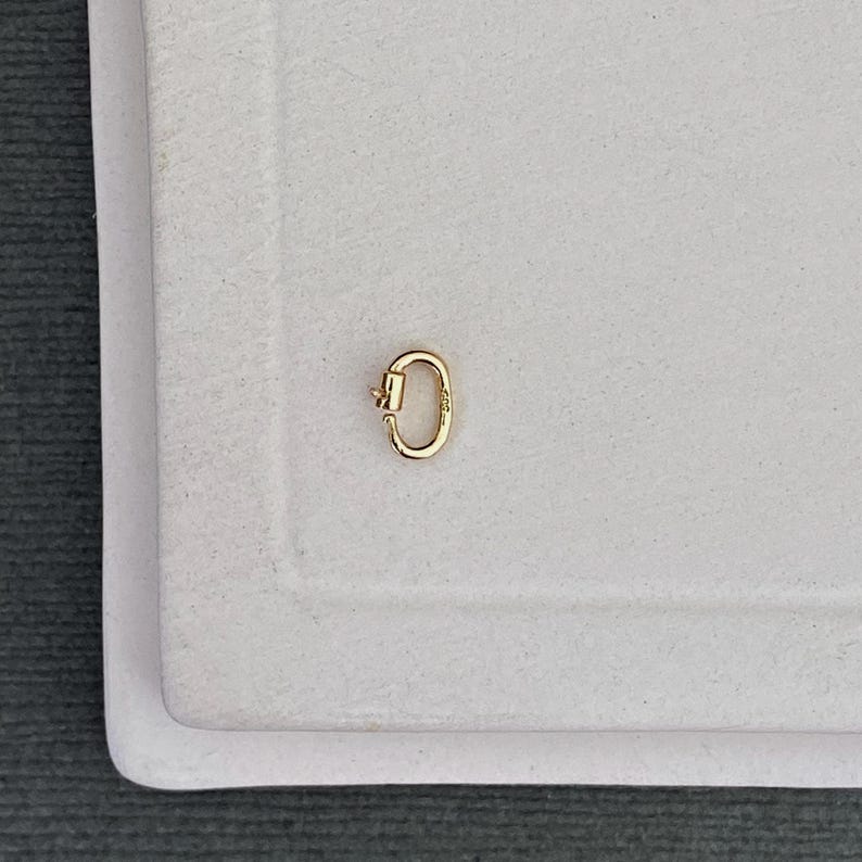 May include: A small, oval-shaped gold carabiner clasp featuring a spring-loaded gate mechanism. The metal component has a polished, reflective surface and is designed for connecting jewellery pieces or charms securely with its functional opening.