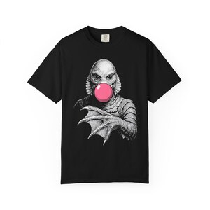 Pode incluir: Camiseta preta com uma ilustração em preto e branco de uma criatura da lagoa negra soprando uma bolha rosa. A criatura tem um rosto de peixe e mãos palmadas. A camiseta é feita de um material macio.