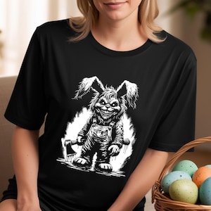 May include: A black t-shirt featuring a white graphic of a creepy bunny rabbit holding an axe. The bunny is wearing overalls and has a menacing grin.