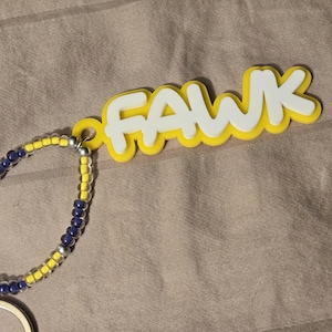 May include: A keychain featuring a white plastic name tag with the text FAWK layered over a yellow plastic backing. The tag is attached to a circular beaded loop made of alternating yellow, dark blue, and silver-colored beads with a metal ring.