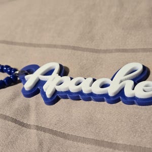 May include: A custom nameplate pendant featuring the word Apache in white script lettering layered over a solid blue background. The pendant is attached to a beaded necklace strand consisting of alternating dark blue and clear light blue beads.