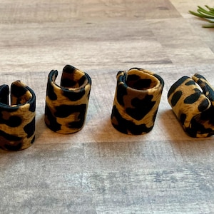 Puede incluir: Seis anillos con estampado de leopardo dispuestos sobre una superficie de madera clara. Los anillos son cilíndricos con una ranura, con un patrón de manchas marrones y negras. El fondo incluye unas ramitas verdes.
