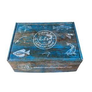 May include: A blue and brown cardboard box with a white logo that says "Fishing & Ed's Outdoor Gear" and a website address, "edsfishingandoutdoorgear.com". The box is decorated with images of fish and fishing lures.