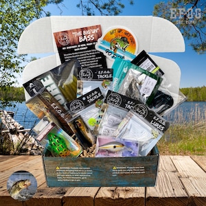May include: An open box filled with fishing lures, tackle, and accessories. The box contains various packaged items, including lures, hooks, and other fishing gear. The box has the text "E.F.O.G. LURES" and "Ed's Fishing Tackle". The background shows a lake and trees.