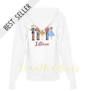 May include: White zip-up hoodie featuring a colorful illustration of nutcracker characters and a ballerina. The word "Lilliana" is written in red script below the illustration. The text "BEST SELLER" is printed on a white diagonal banner.
