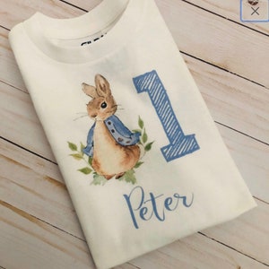 May include: White t-shirt featuring a watercolor illustration of Peter Rabbit wearing a blue jacket, with the number 1 in blue and the name "Peter" in script. The design is centered on the front of the shirt.