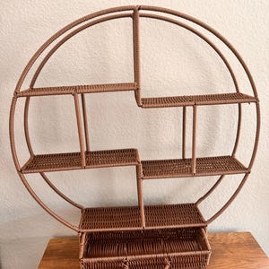 Pode incluir: Uma prateleira de parede redonda marrom com v&aacute;rias prateleiras tecidas e uma gaveta. A prateleira tem um design exclusivo com uma grande moldura circular e v&aacute;rias prateleiras retangulares de tamanhos variados. A gaveta tem uma al&ccedil;a para f&aacute;cil acesso.