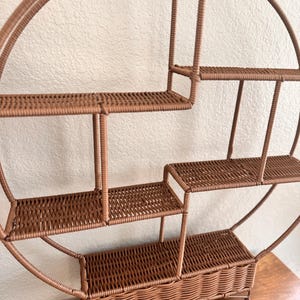 Pode incluir: Uma prateleira de parede redonda marrom com v&aacute;rias prateleiras tecidas e uma base em forma de cesta. A prateleira tem um design em camadas com tamanhos de prateleiras vari&aacute;veis, perfeita para exibir itens pequenos. A estrutura &eacute; feita de um material resistente.
