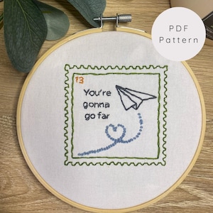 May include: Embroidered hoop art with a green and white frame. The design is a white paper airplane with a blue heart and trail, and the text "You're gonna go far" in black.