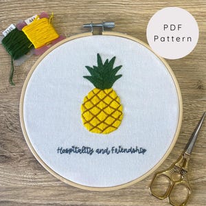 Peut inclure: Broderie d'ananas dans un cercle en bois. L'ananas est jaune et vert, avec le texte "Hospitality and Friendship" en dessous. Comprend des bobines de fil vert et jaune et une paire de ciseaux dorés. Un cercle blanc avec "PDF Pattern" est en haut à droite.