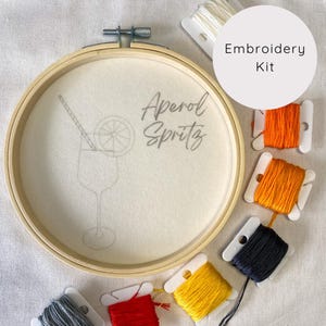 May include: Embroidery kit with a hoop featuring a gray outline of a cocktail glass with a lemon slice and straw, and the text "Aperol Spritz". The kit includes five spools of embroidery floss in orange, yellow, red, black, and gray.