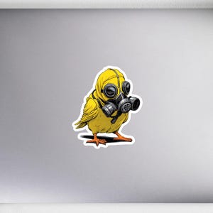 May include: A yellow sticker featuring a cartoon bird wearing a yellow hazmat suit and a black gas mask. The bird is standing on a white surface. The sticker is on a laptop with a notebook, pencils, headphones, a plant, and a mouse in the background.