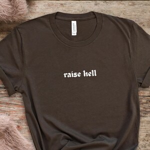 May include: A dark brown t-shirt with the phrase "raise hell" printed in white, gothic-style lettering. The shirt has a crew neck and short sleeves. The brand "Canvas" is printed on the upper right side of the shirt.
