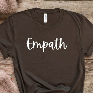Puede incluir: Una camiseta marrón oscuro con la palabra "Empath" impresa en blanco en la parte delantera.