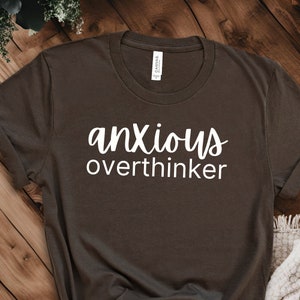 Könnte beinhalten: Ein dunkelbraunes T-Shirt mit den Worten "anxious overthinker" in weißer Schrift. Das T-Shirt hat einen Rundhalsausschnitt und besteht aus einem weichen Material. Das T-Shirt wird auf einer Holzoberfläche präsentiert.