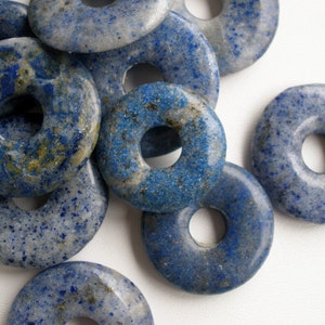 May include: A collection of blue lapis lazuli donut-shaped beads. The beads have a speckled appearance with varying shades of blue and gray. Each bead has a hole in the center. The beads are suitable for jewelry making.
