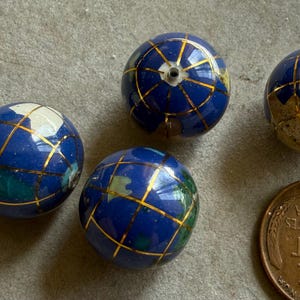 May include: Four blue globe-shaped beads with gold grid lines and continents. The beads are next to a US one-cent coin. The beads are a mix of blue, white, and green. The beads are likely for jewelry making or crafts.