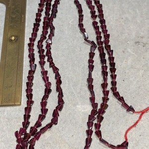May include: Multiple strands of dark red, triangular beads. The beads are strung together, forming necklaces. A brass ruler is in the background, with red thread and tassels. The beads are likely made of glass.