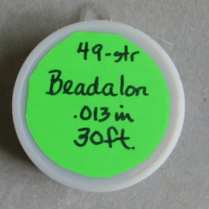 May include: A green label with black text on a white plastic container. The label reads "49-str Beadalon .013 in 30ft."