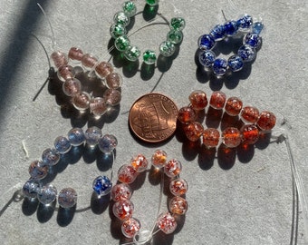 May include: Six strands of glass beads, each with a different color and pattern. The beads are strung on clear elastic cord. The beads are all round and have a speckled appearance. The beads are arranged in a circle on a gray surface.