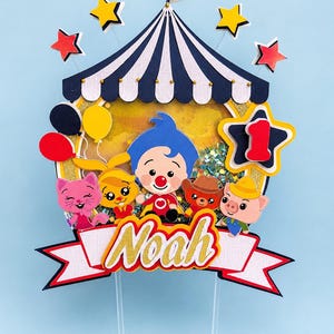 May include: A colorful circus-themed cake topper with a blue and white striped tent, stars, balloons, and cartoon characters. The topper includes the name "Noah" in gold glitter lettering and a number one star.