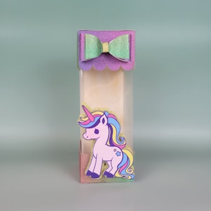 May include: A small, clear box with a purple and green glitter bow on top. Inside the box is a pink unicorn with a rainbow mane and tail.