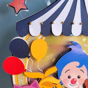 May include: A colorful circus-themed decoration featuring a blue and white striped tent top, balloons, and cartoon characters. Includes a red, yellow, and blue color scheme with gold accents. The design includes a pink cat, a yellow bunny, and a clown.