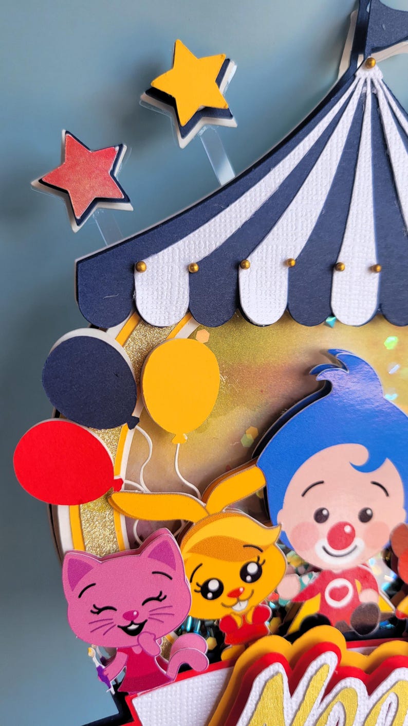 May include: A colorful circus-themed decoration featuring a blue and white striped tent top, balloons, and cartoon characters. Includes a red, yellow, and blue color scheme with gold accents. The design includes a pink cat, a yellow bunny, and a clown.
