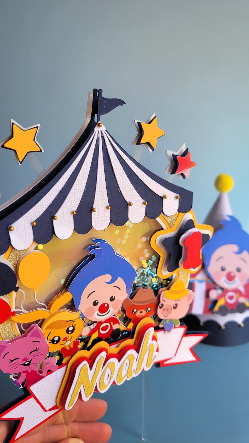May include: A colorful circus-themed cake topper with a blue and white striped tent, stars, and cartoon characters. The topper includes the name "Noah" and the number "1", perfect for a first birthday celebration.