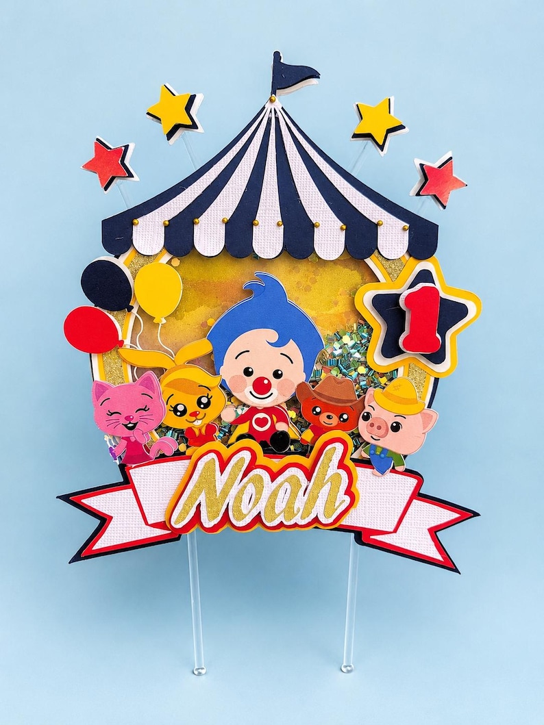 May include: A colorful circus-themed cake topper with a blue and white striped tent, stars, balloons, and cartoon characters. The topper includes the name "Noah" in gold glitter lettering and a number one star.