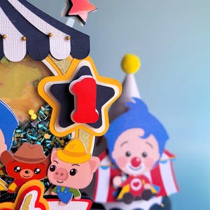 May include: A colorful circus-themed cake topper with a star-shaped number one, a clown, and cartoon animal characters. The topper features a circus tent design with a blue, white, and gold color scheme, perfect for a first birthday celebration.