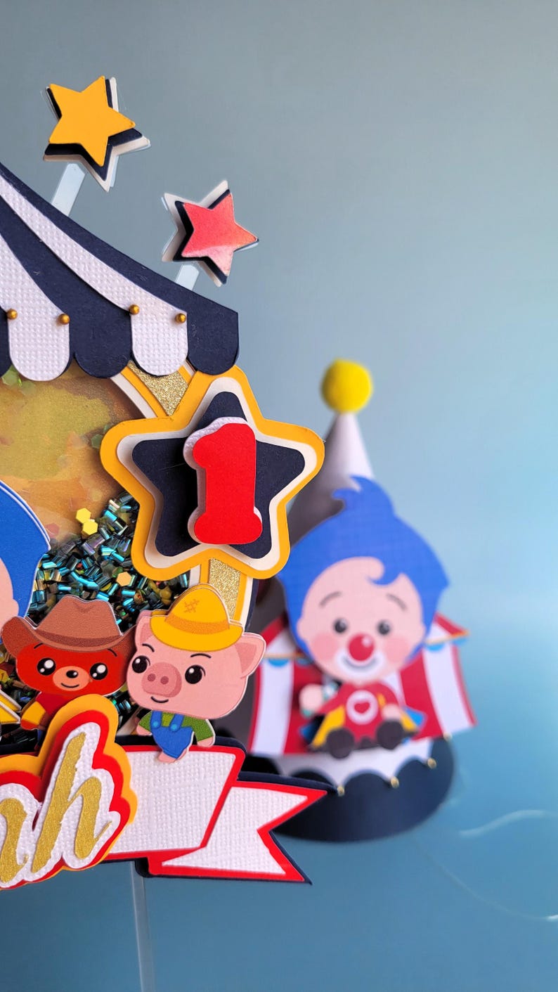 May include: A colorful circus-themed cake topper with a star-shaped number one, a clown, and cartoon animal characters. The topper features a circus tent design with a blue, white, and gold color scheme, perfect for a first birthday celebration.