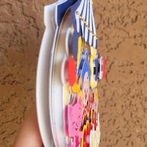 May include: A colorful, layered circus-themed decoration. The design features a circus tent, stars, and cartoon characters in shades of blue, red, yellow, and pink. The decoration is held by a hand.