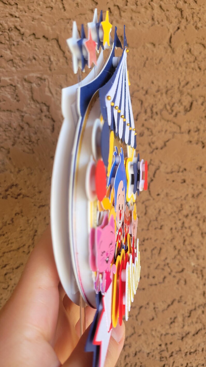 May include: A colorful, layered circus-themed decoration. The design features a circus tent, stars, and cartoon characters in shades of blue, red, yellow, and pink. The decoration is held by a hand.