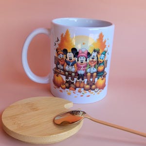 May include: A white ceramic mug featuring a colorful illustration of cartoon characters sitting on a bench with pumpkins and autumn leaves. The mug includes a round wooden lid and a gold-toned metal spoon resting on the lid.