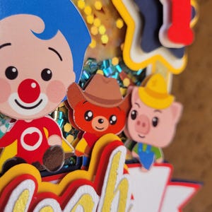 May include: A colorful birthday cake topper featuring cartoon characters, including a clown with blue hair, a bear in a cowboy hat, and a pig. The name "Noah" is prominently displayed in gold lettering.