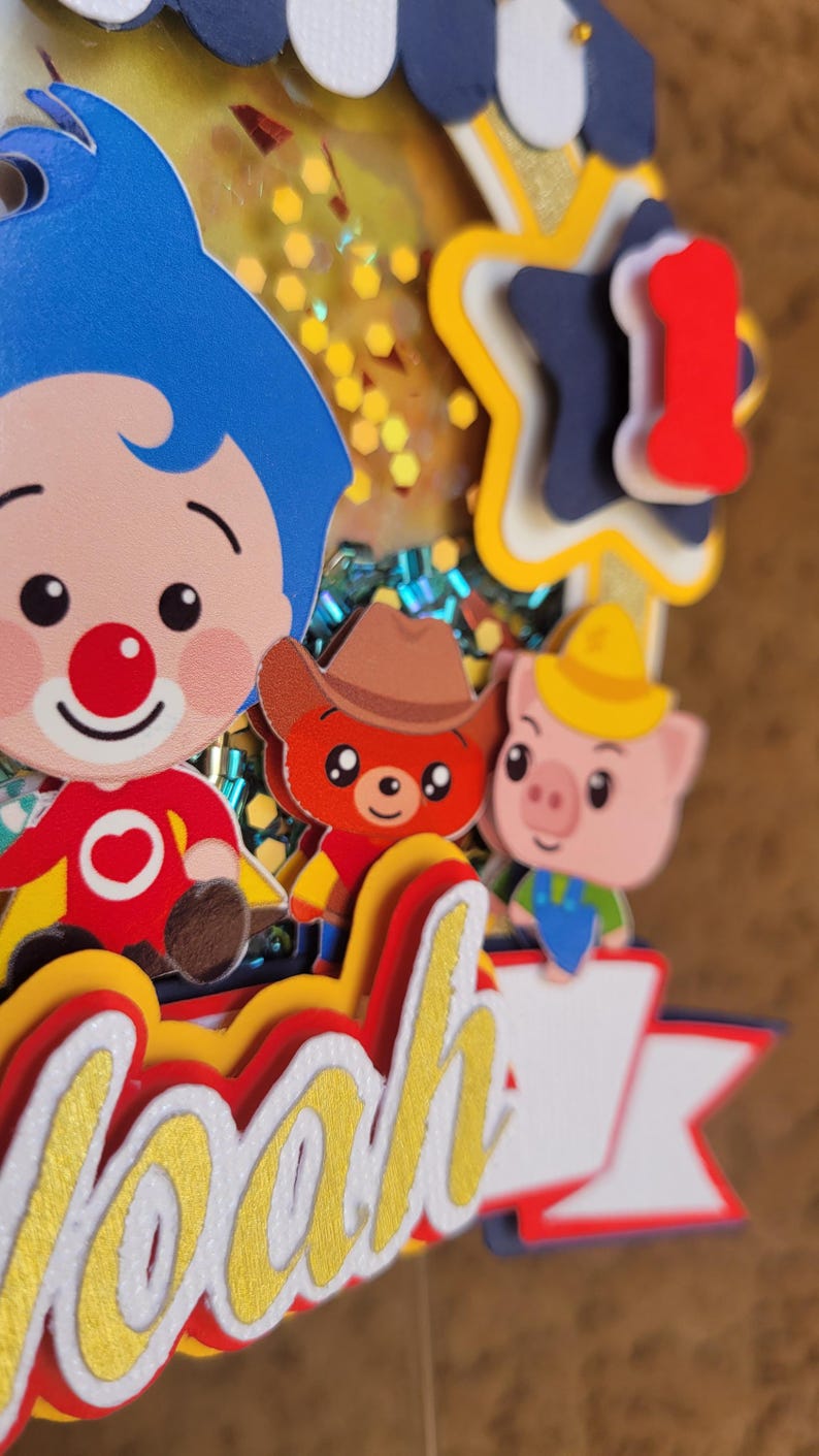 May include: A colorful birthday cake topper featuring cartoon characters, including a clown with blue hair, a bear in a cowboy hat, and a pig. The name "Noah" is prominently displayed in gold lettering.