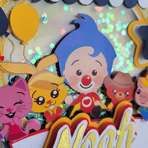 May include: A colorful, layered cake topper featuring cartoon characters and the name "Noah" in gold lettering. The design includes a circus tent, balloons, and various animal figures, creating a festive and celebratory theme.